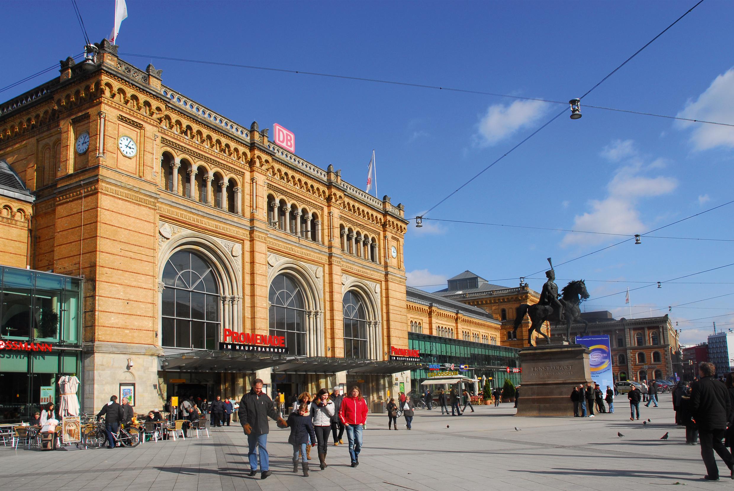 People walk, stand or sit on chairs in front of the station building of Hannover Hauptbahnhof.