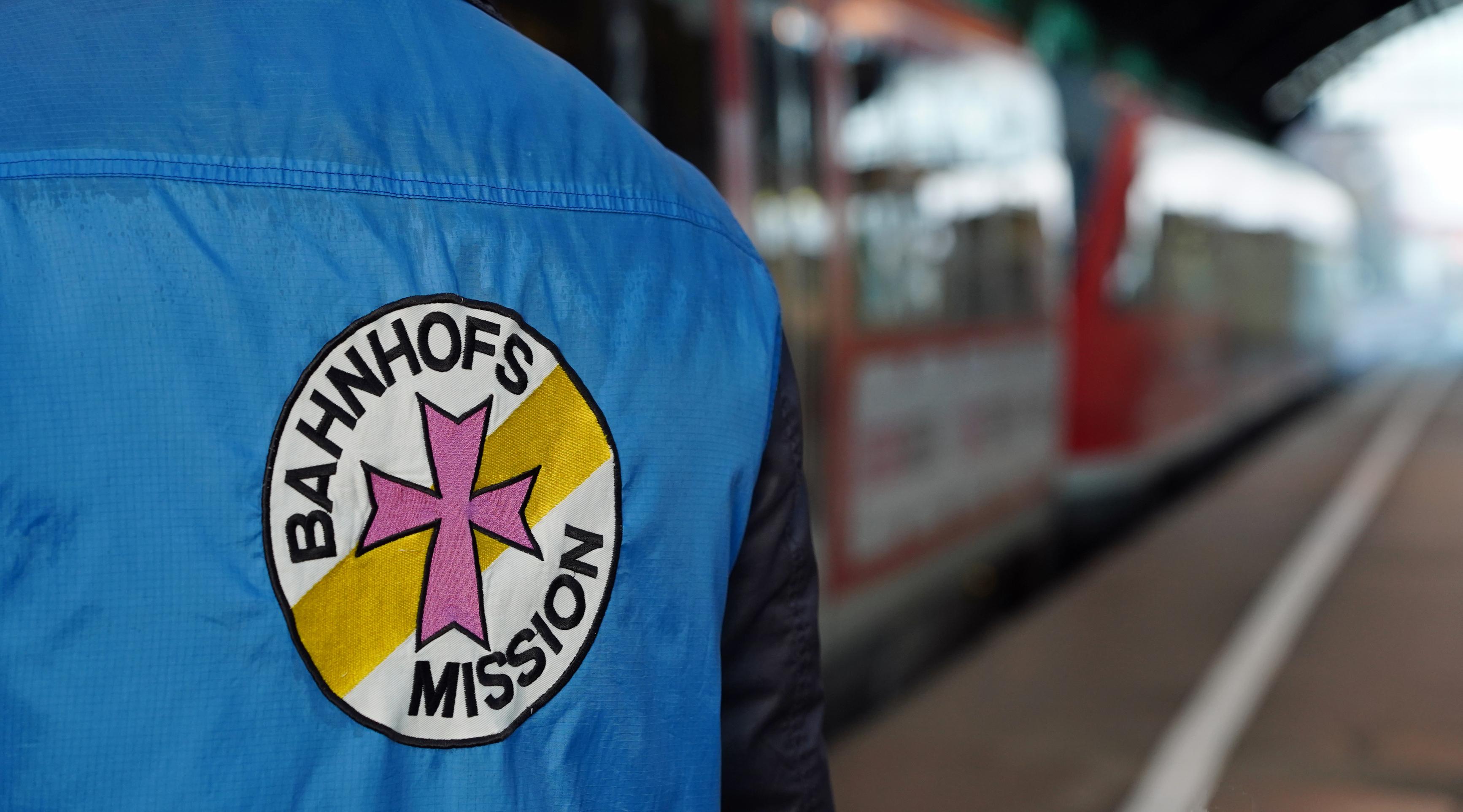 Eine Person am Gleis trägt eine Weste, auf deren Rückseite sich das Logo der Bahnhofsmission befindet.