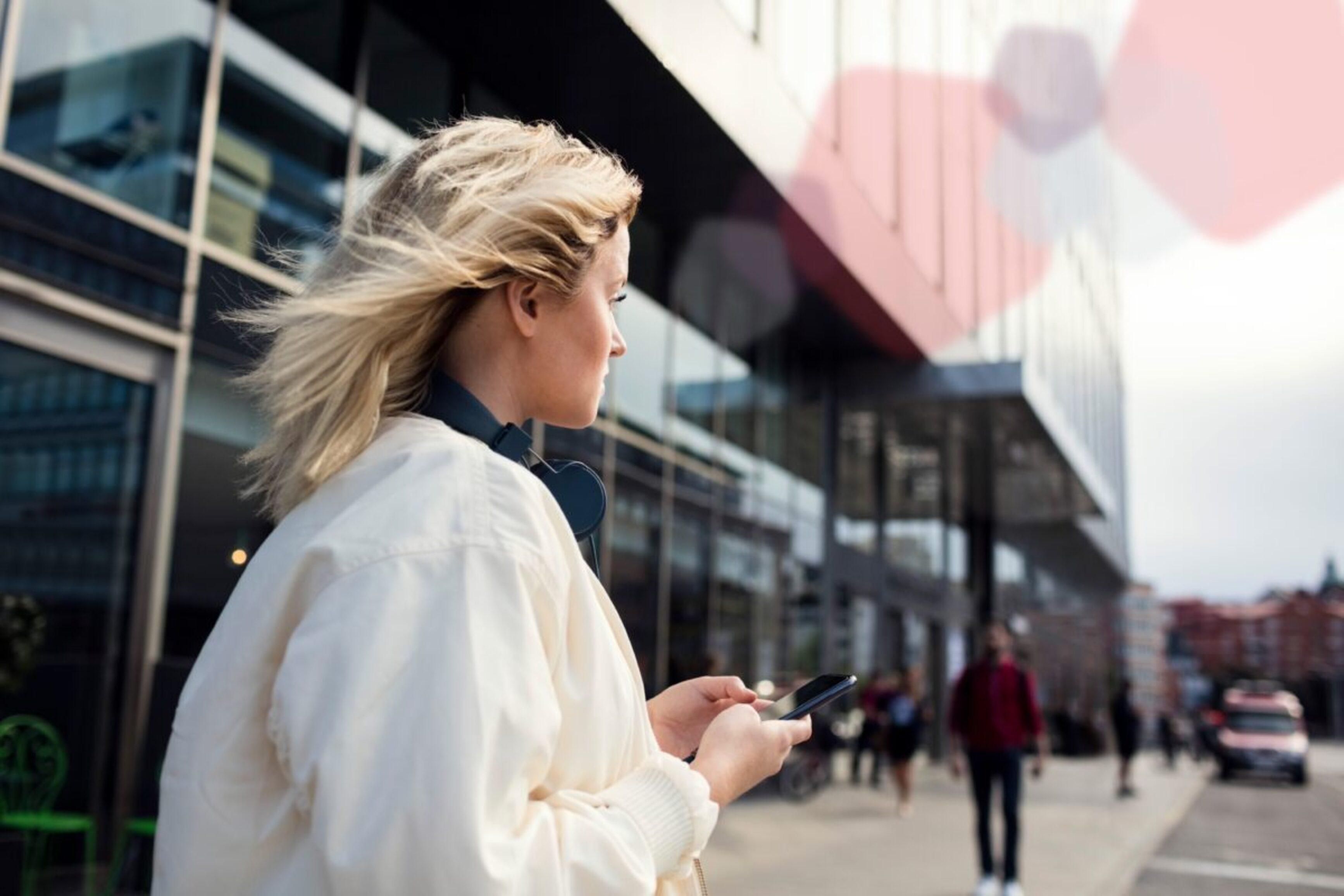 A woman stands with her smartphone in front of a train station and looks into the distance.