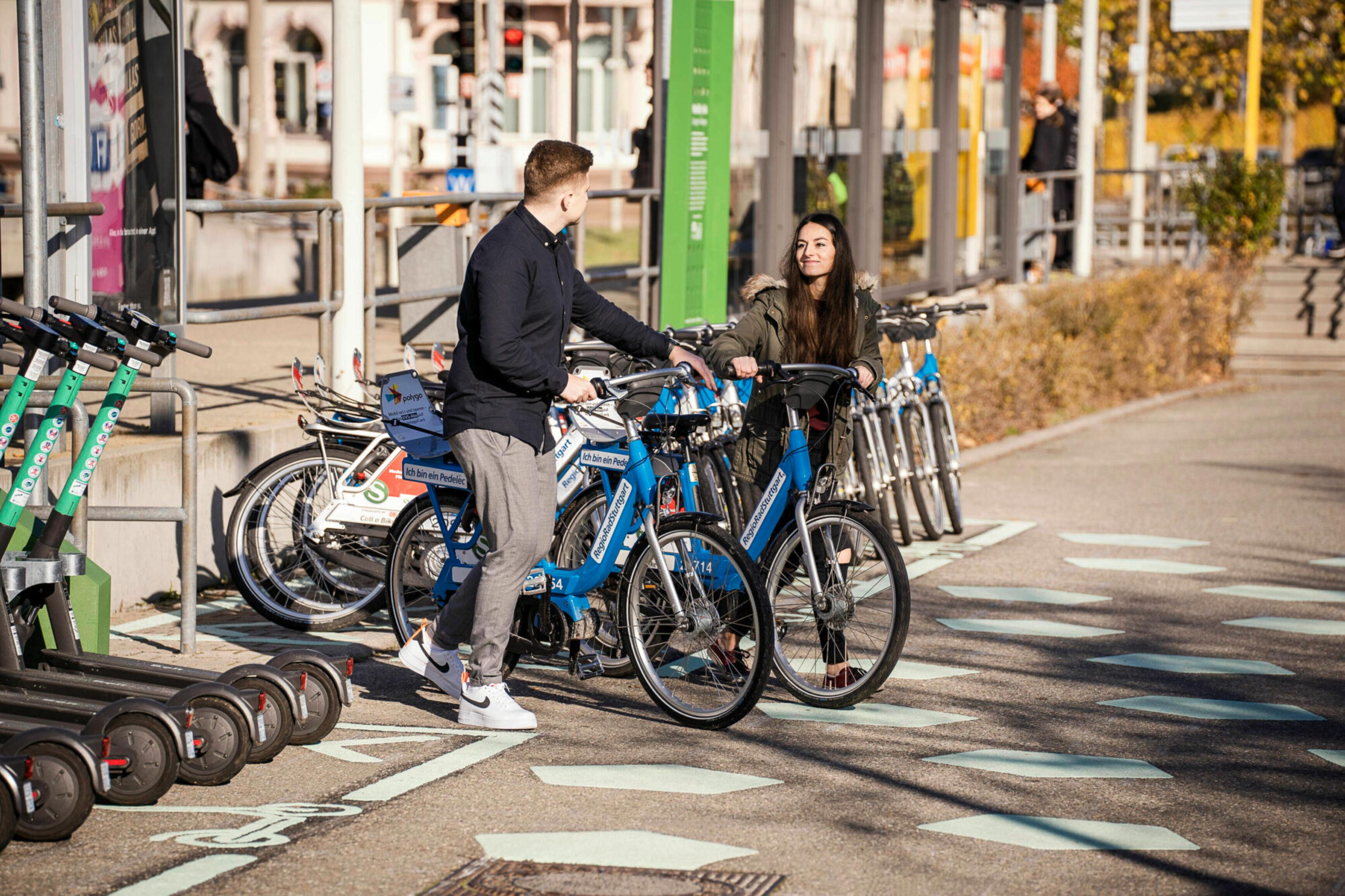 A woman and a man stand with two blue bicycles at a Mobility Hub next to a railway station.