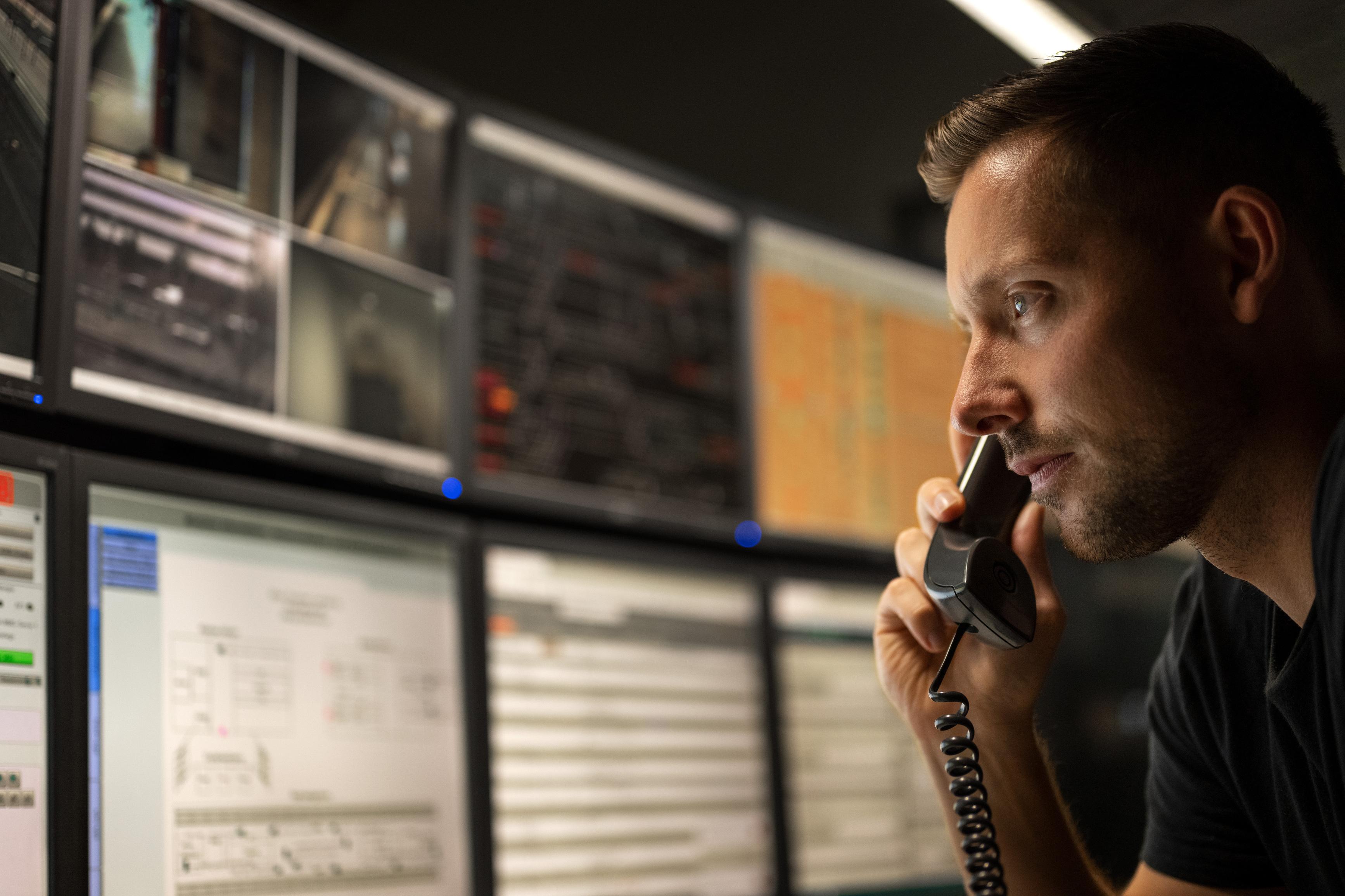 An employee on the phone in the 3-S centre in front of a row of monitors.