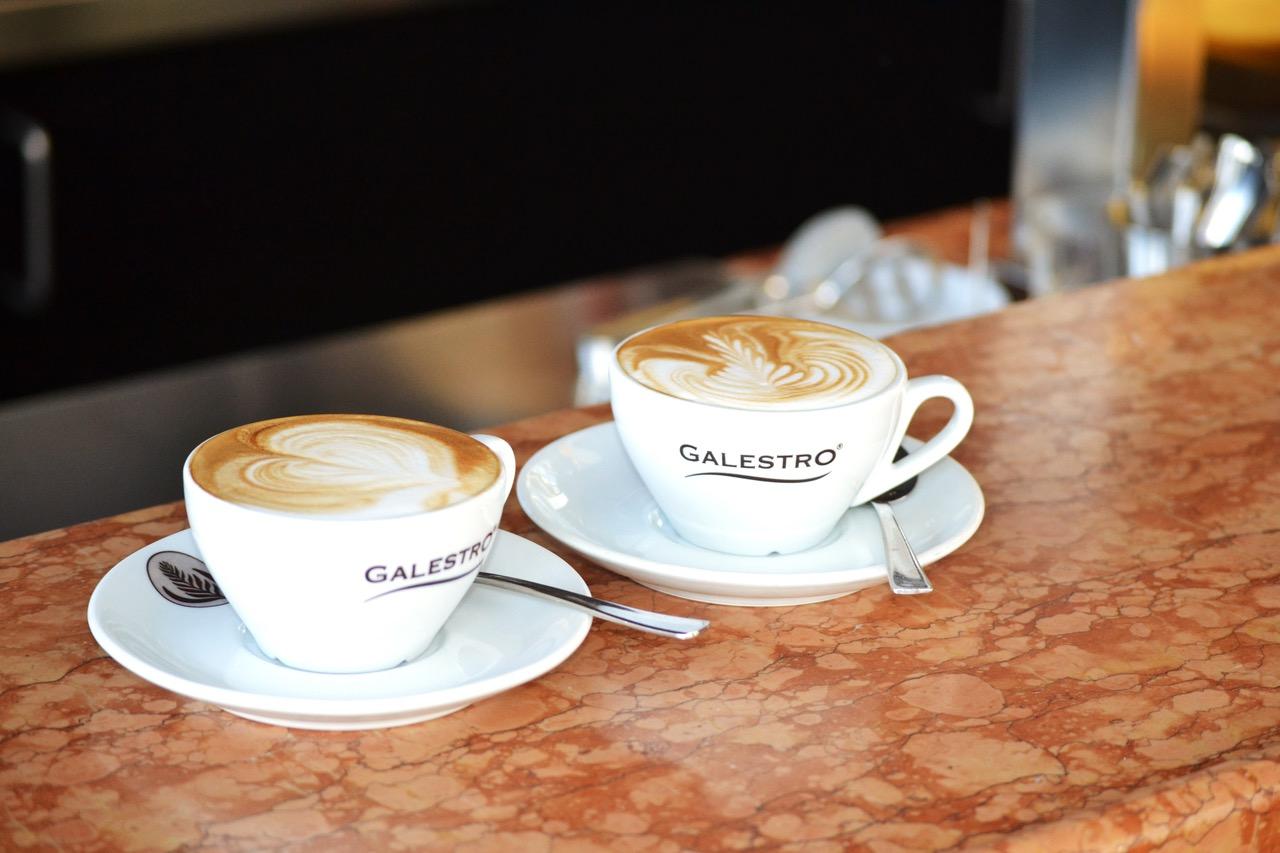 Zwei volle Cappuccino-Tassen mit Galestro-Schriftzug stehen auf einem bräunlich gemusterten Tresen.